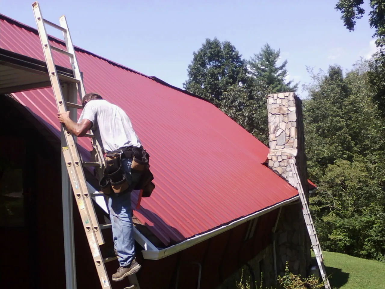 Roofer on a ladder beside a red metal roof and stone chimney — Soffit & Fascia Repair by Roofing Brothers in Beltrami